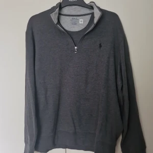 Mörkgrå half zip tröja Polo Ralph Lauren - Mörkgrå långärmad tröja från Polo Ralph Lauren med klassisk broderad logga på bröstet. Tröjan har en snygg half zip-dragkedja och hög krage. Tillverkad i mjuk bomullsmix, perfekt för lager-på-lager och chill stil.