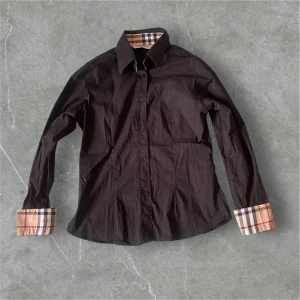 Svart Burberry skjorta - Snygg svart skjorta från Burberry med klassiska rutiga detaljer på insidan av kragen och ärmsluten. Skjortan har knappar framtill, lång ärm och diskret broderad logga på bröstet. Tillverkad i bomull för en skön känsla och stilren look.