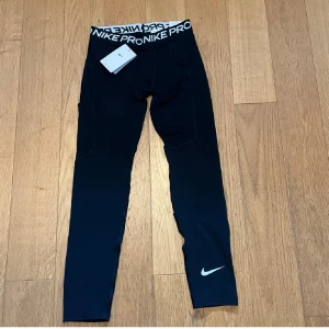 Nike Pro svarta tights  - Svarta Nike Pro tights med vit logga på benet och bred resår i midjan med Nike Pro-tryck. Tillverkade i Dri-FIT-material som andas och transporterar bort fukt. Perfekta för träning och sportiga aktiviteter.perfelt för löpturer eller till sport som basket.      Pris kan diskuteras    Storlek XL junior (xs/ 164-170)