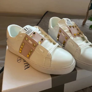 Valentino Rockstud vita sneakers - Snygga vita Valentino Rockstud sneakers med ljusrosa detaljer och guldiga nitar. Skorna har rund tå, platt sula och är tillverkade i skinn. Klassisk och lyxig design med snörning och ikoniska rockstuds längs sidorna.