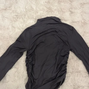 Mörkgrå långärmad topp med rynkade detaljer - Säljer en mörkgrå långärmad topp med snygga rynkade detaljer längs sidorna. Tunt och mjukt material som sitter skönt på kroppen. Perfekt för dig som gillar en stilren men ändå trendig look.