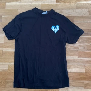 Svart t-shirt med blått hjärta från Weekday - Svart t-shirt från Weekday med ett tryckt blått hjärta på bröstet. Klassisk passform med rund hals och korta ärmar. 