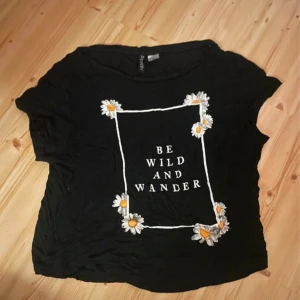 Svart t-shirt med prästkragar från Divided - Svart t-shirt från Divided med tryck av vita prästkragar och texten 'Be Wild And Wander' på framsidan. T-shirten har korta ärmar och en avslappnad passform. Perfekt för en cool och avslappnad stil.