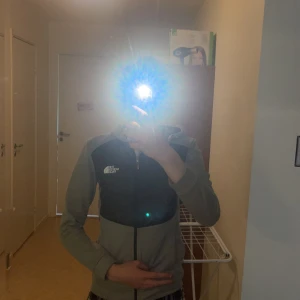 Grå och svart cardiganhoodie från The North Face - Säljer en grå och svart cardigan från The North Face med dragkedja framtill. Den har lång ärm och en sportig look, perfekt för dig som gillar streetwear. Loggan syns tydligt på bröstet och jackan har en bekväm passform.