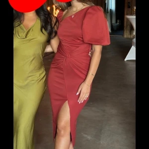 Rostfärgad klänning med puffärm och slits - Säljer en rostfärgad klänning med markerad midja, puffärmar och snygg slits framtill. Klänningen har en twistad detalj vid midjan och är figurnära med en elegant v-ringning. Perfekt för festliga tillfällen.