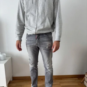 Grå jeans från Dsquared2 - Säljer ett par grå jeans från Dsquared2 i modell Cool Guy. Byxorna har slitna detaljer, dragkedja och knappar, samt en svart '2' på ena benet. Klassisk femficksmodell med normal passform och snygg tvätt. Sitter som storlek 48! Men är storlek 46!