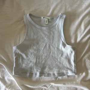 Ljusgrå croppad ribbad linnetopp från H&M - Säljer en ljusgrå croppad linnetopp från H&M. Toppen är ribbad och har en ärmlös design med rund halsringning. Perfekt för varma dagar eller som lager under en skjorta.