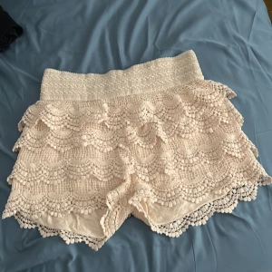 Vita virkade shorts med volang - Säljer ett par vita virkade shorts med bred resår i midjan och flera lager volangdetaljer. De har en feminin och bohemisk stil med fint spetsmönster och scallop-kant nedtill. Perfekta för sommardagar!
