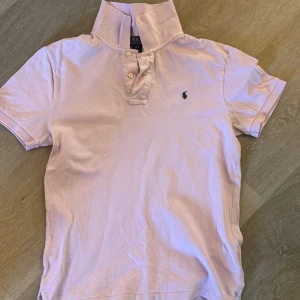rosa pikét från Polo Ralph Lauren - Säljer en klassisk ljusrosa pikétröja från Polo Ralph Lauren med broderad logga på bröstet. Tröjan har korta ärmar, krage och knappar vid halsen. Perfekt för en stilren och avslappnad look.