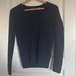 Mörkblå stickad tröja från Ralph Lauren -  HELT NY OANDVÄND Säljer en klassisk mörkblå stickad tröja från Ralph Lauren med v-ringning och diskret röd logga broderad på bröstet. Tröjan har lång ärm och smal passform, perfekt till jeans eller kjol.