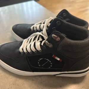 Svarta sneakers från Pier One med högt skaft nya med prislapp - Säljer ett par svarta sneakers från Pier One med högt skaft och vita snören. Skorna har vita sulor, metallhål för snörningen och dekorativa detaljer på sidan. Perfekta för en stilren och avslappnad look.