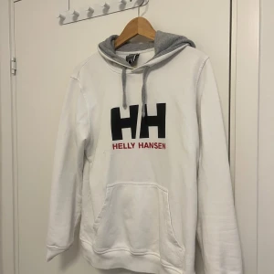 Vit hoodie från Helly Hansen - Vit hoodie från Helly Hansen med stor svart HH-logga och röd text på bröstet. Grå insida på huvan och gråa snören. Klassisk känguruficka framtill och mjukt material. Perfekt för en avslappnad stil.