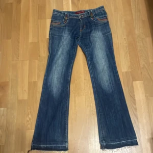 Blå bootcut jeans med broderade detaljer - Säljer ett par blå bootcut jeans med snygga slitningar och broderade detaljer på bakfickorna och är dessutom low waist. Jeansen har normal passform och klassisk femficksdesign med knappar och dragkedja. Perfekta för dig som gillar en avslappnad men ändå trendig stil. Jeansen är dock korta, jag är 165 och dom är korta för mig så dessa passar perfekt för dig som är kortare.