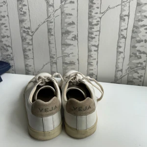 Vita sneakers från Veja med beige detaljer - Säljer ett par klassiska vita sneakers från Veja med beige mockadetaljer vid hälen och vita snören. De går säkert att tvätta av de för att få en fräschare look. 