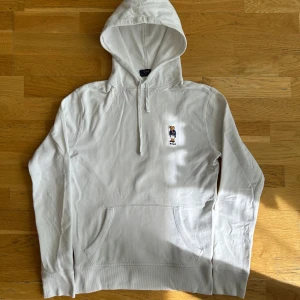 Vit Polo Ralph Lauren hoodie  - Vit hoodie från Polo Ralph Lauren med broderad björn på bröstet. Tröjan har huva med dragsko och en magficka.