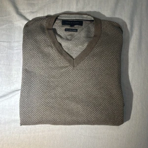Beige v-ringad tröja från Tommy Hilfiger - Säljer en beige v-ringad tröja från Tommy Hilfiger i Pima Cotton Cashmere. Tröjan har ett diskret fiskbensmönster och ribbade muddar vid ärmslut och nederkant. Perfekt för lager-på-lager och stilrena outfits. Super skönt material