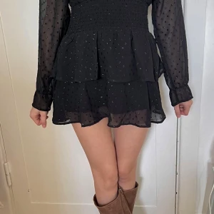 Svart kortklänning  - Supersöt svart kortklänning med volanger och diskret prickigt mönster. Klänningen är långärmad och smock i midjan för en snygg passform. Perfekt för dig som gillar en feminin och trendig stil💕