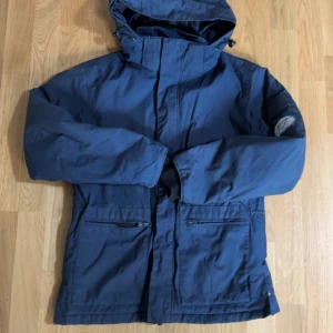 Mörkblå vindjacka med huva - Säljer en mörkblå vindjacka med huva och dragkedja framtill. Jackan har två framfickor med dragkedja och justerbar huva. Perfekt för blåsiga dagar och enkel att matcha med olika outfits.