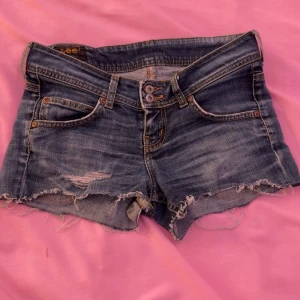 Blå jeansshorts från Lee med fransar - Säljer ett par blå jeansshorts från Lee med råa fransar nertill och slitningar framtill. Klassisk femficksmodell med dubbla knappar i midjan och Lee-logga bak. Perfekta för sommaren!