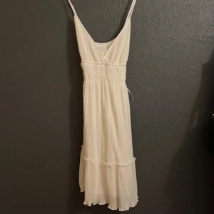 Vit plisserad klänning från Gina Tricot - Säljer en vit plisserad klänning från Gina Tricot med smala axelband och v-ringning. Klänningen har markerad midja och volangdetalj nertill, perfekt för sommarens alla tillfällen. Lätt och luftig modell med fin struktur.