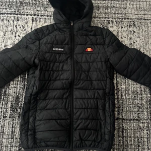 Svart quiltad pufferjacka från Ellesse - Säljer en svart pufferjacka från Ellesse med huva och dragkedja framtill. Jackan har quiltat mönster och Ellesse-logga på bröstet. Perfekt för kyliga dagar och enkel att matcha med olika outfits. Pris kan såklart diskuteras men kan inte gå ner för mycket!