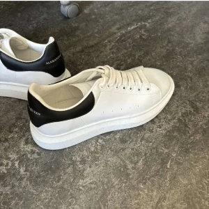 Vita sneakers från Alexander McQueen - Säljer ett par klassiska vita sneakers från Alexander McQueen med svart hälparti och tjock vit sula. Skorna har snörning och en stilren design som passar till det mesta. Perfekta för dig som gillar trendiga och bekväma skor.