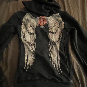 Svart hoodie med vingar från Sinful - Säljer en svart hoodie från Sinful med dragkedja och huva. På ryggen finns ett stort tryck med glittriga änglavingar och ett rött S. Framtill står det Sinful på bröstet. Perfekt för dig som gillar unika detaljer!