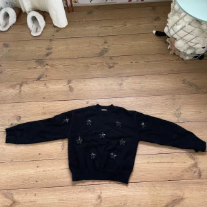 Svart stickad tröja med stjärnor - Svart stickad tröja med rund halsringning och långa ärmar. Tröjan har dekorativa stjärnor framtill i ett glittrigt material som ger en cool touch. Perfekt för dig som vill ha något unikt och snyggt i garderoben.