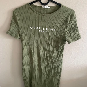 Olivgrön ribbad klänning med tryck - Säljer en olivgrön ribbad klänning med texten 'C'est la vie Paris' framtill. Klänningen har korta ärmar och rund halsringning. Perfekt för en avslappnad stil.