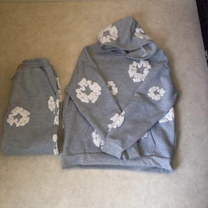 Grå hoodie från Denim Tears med vita blommor - Säljer en grå hoodie från Denim Tears med vita blommönster tryckta över hela tröjan. Hoodien har en klassisk passform med huva och ribbade muddar. Perfekt för dig som vill ha något unikt och stilrent.