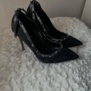 Svarta pumps med nitar från Juliet - Snygga svarta pumps från Juliet med spetsig tå och klack. Skorna har dekorativa silverfärgade nitar längs kanterna och remmar runt vristen. 
