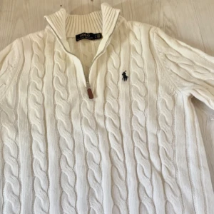Polo Ralph Lauren || Vit Stickad Quarterzip - Säljer en klassisk vit kabelstickad tröja från Polo Ralph Lauren med halv dragkedja och broderad logga på bröstet. Tröjan har lång ärm, hög krage och ribbade muddar. Perfekt för en stilren och tidlös look.
