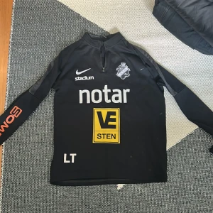 Svart AIK fotbollströja från Nike - Säljer en svart långärmad AIK fotbollströja från Nike med dragkedja vid halsen. Tröjan har flera sponsorer tryckta på både fram- och baksidan, bland annat notar, VE Sten och SVEA. AIK:s klubbmärke finns på bröstet och materialet är Dri-Fit.