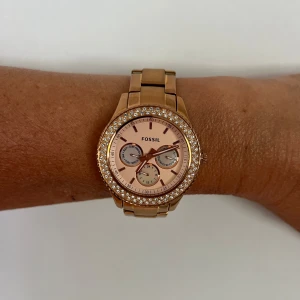 Guldig klocka från Fossil med glittrig kant - Perfekt till studenten/skolavslutningen. Säljer en snygg guldig klocka från Fossil med glittrig stenkant runt urtavlan. Klockan har en rund form, länkarmband och flera små urtavlor på insidan. Perfekt accessoar för att lyfta din stil. Nytt batteri bytades förra veckan.