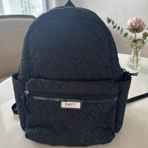 Marlene Birger DAY Svart quiltad ryggsäck  - Säljer en svart quiltad ryggsäck från DAY med flera fack och dragkedja framtill. Väskan har vadderade axelremmar och sidofickor, perfekt för att bära med sig allt man behöver. Klassisk och stilren design med mönstrad yta.