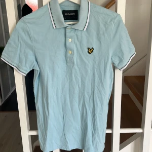 Ljusblå pikétröja från Lyle & Scott - Säljer en ljusblå pikétröja från Lyle & Scott med klassisk krage och två knappar framtill. Tröjan har vit och mörkblå rand på krage och ärmslut samt den ikoniska gula loggan broderad på bröstet.
