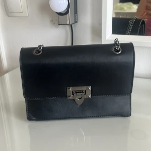 Svart axelväska med kedja - Snygg svart axelväska med silverfärgad kedjerem och metallspänne framtill. Väskan har en stilren, rektangulär form och är tillverkad i ett slätt material som liknar skinn. Perfekt för att bära det viktigaste.