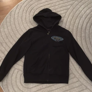 Svart hoodie från Ed Hardy med tryck - Svart hoodie från Ed Hardy med dragkedja och huva. Tryck på bröstet med texten 'Ed Hardy Tattoo The World Est 1971' och stort färgglatt motiv samt Ed Hardy-logga på ryggen. Perfekt för dig som gillar streetwear och unika detaljer.