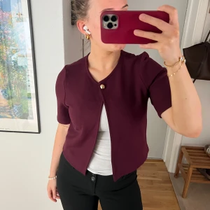 Vinröd kortärmad kofta från Gina Tricot - Säljer en vinröd kortärmad kofta från Gina Tricot med en dekorativ knapp framtill. Perfekt att ha över ett linne eller t-shirt. Enkel och stilren design som passar till många outfits.