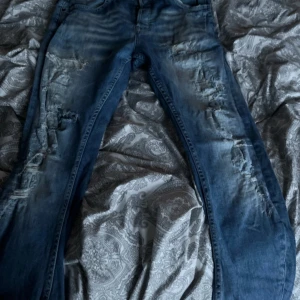 Blå slitna jeans med trasiga detaljer - Säljer ett par blå jeans med flera slitningar och trasiga detaljer på både fram- och baksida. Jeansen har klassisk femficksmodell och normal passform. Perfekta för en avslappnad och trendig look.