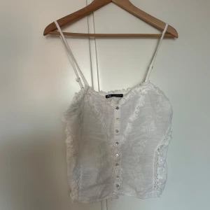 Vit linnetopp med spetsdetaljer från Zara - Säljer en söt vit linnetopp från Zara med smala axelband, knappar framtill och fina spetsdetaljer längs kanterna. Toppen har ett diskret blommönster i tyget och är perfekt för varma dagar.