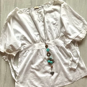 Vit blus med accessoar - Vit blus med bred midja och knytband, puffiga trekvartsärmar och v-ringad hals. Lätt och luftig modell, perfekt för varmare dagar. Snygg att styla med smycken för en personlig touch.