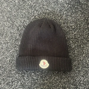 Svart ribbad mössa från Moncler - Svart ribbstickad mössa från Moncler med uppvikt kant och klassisk Moncler-logga framtill. Perfekt för kyliga dagar och enkel att matcha med olika outfits.
