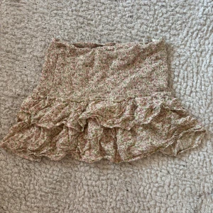 Blommig volangkjol från Petits - Supersöt kort blommig kjol från Petits med volanger och smockad midja. Kjolen har en ljus bas med små gröna och rosa blommor, perfekt för en somrig look. Lätt och luftig modell som är enkel att styla med en enkel topp.