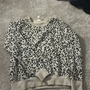 Beige leopardmönstrad sweatshirt från H&M - Säljer en beige sweatshirt från H&M med svart leopardmönster. Tröjan har rund halsringning, ribbade muddar vid ärmslut och nederkant samt lång ärm. Perfekt för en trendig och avslappnad stil.