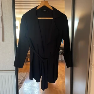 Svart trenchcoat med bälte från BikBok - Säljer en svart trenchcoat från BikBok i storlek S. Jackan har lång ärm, klassisk krage och knytbälte i midjan för en snygg siluett. Perfekt för övergångssäsonger och enkel att matcha med olika outfits.