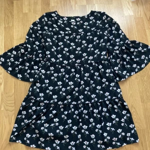 Svart blommig klänning från GAP - Supersöt svart klänning från GAP med vitt blommönster och trekvartsärmar. Klänningen har volangdetaljer och knyt i midjan för en fin passform. Perfekt för dig som gillar en feminin och bekväm stil.