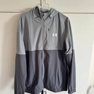 Grå vindjacka från Under Armour - Snygg tvåfärgad grå vindjacka från Under Armour med huva och dragkedja. Jackan har en sportig look och en liten logga på bröstet. Perfekt för utomhusaktiviteter och enkel att matcha med andra plagg.