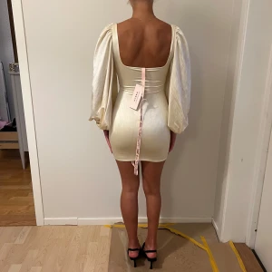 Oanvänd OhPolly Champagnefärgad klänning med puffärmar - Supersnygg kort klänning från OhPolly i champagnefärgad satin med puffiga långärmar och cut-out framtill. Aldrig använd. Köptes för 600 kr.