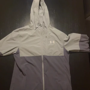 Grå vindjacka från Under Armour - Snygg tvåfärgad vindjacka från Under Armour i ljusgrått och mörkgrått. Jackan har huva med dragsko, hel dragkedja framtill och diskret logga på bröstet. Perfekt för blåsiga dagar och sportiga outfits.